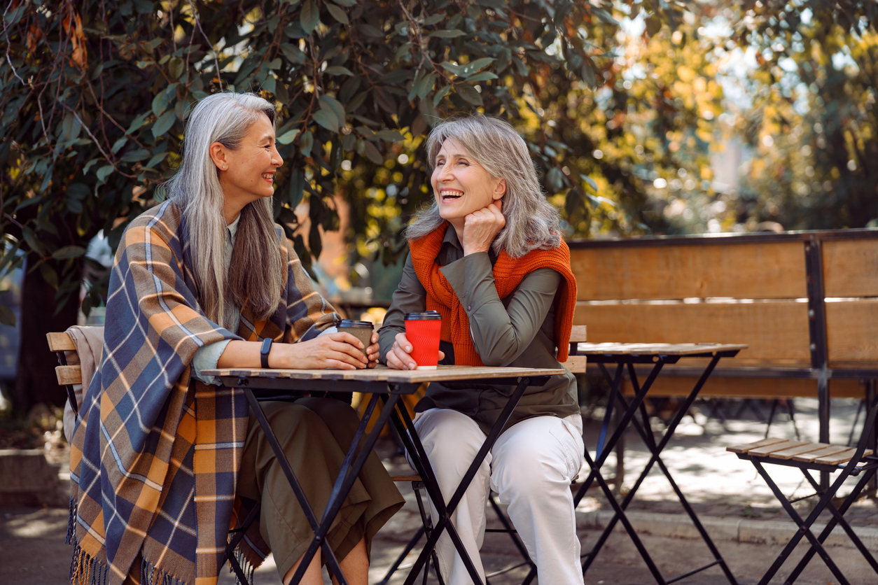 Mature Asian lady with positive grey haired friend spend time together in street cafe